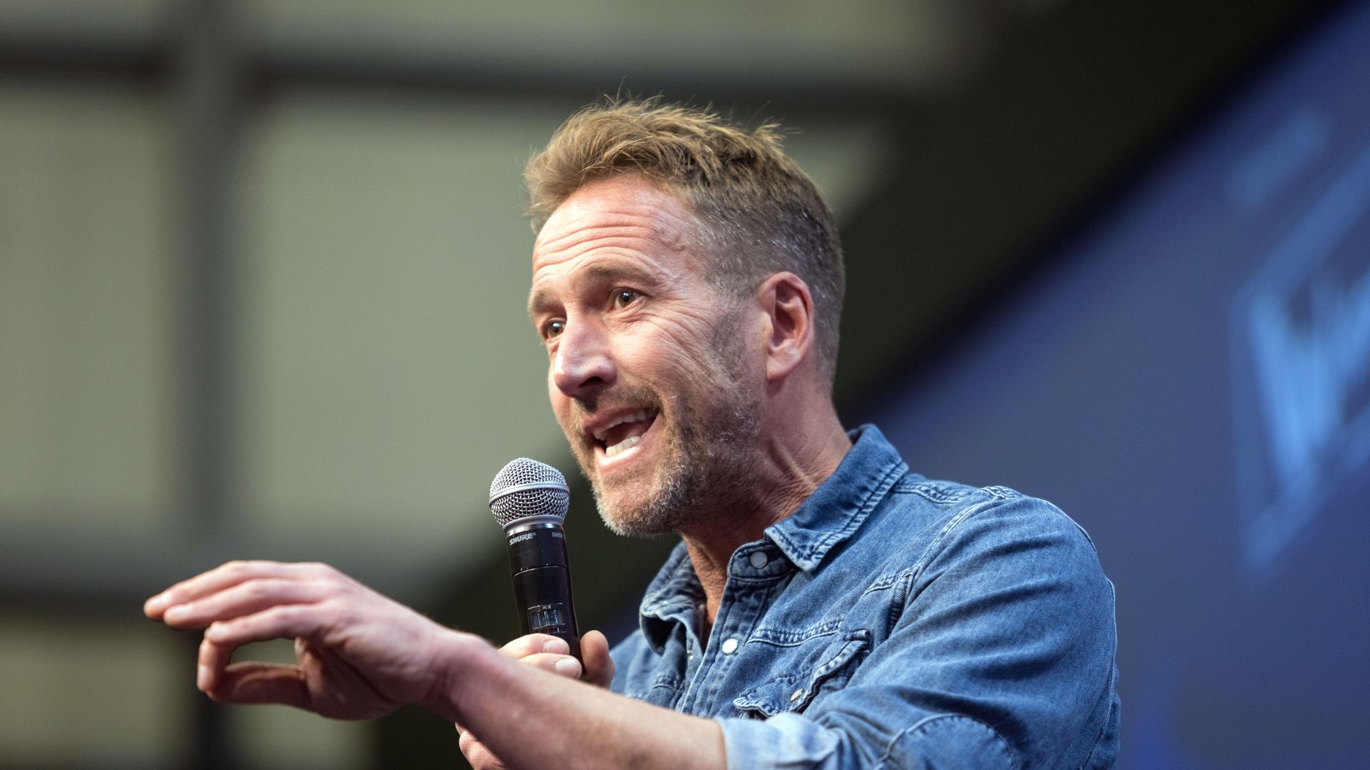 man in denim shirt speaking on stage