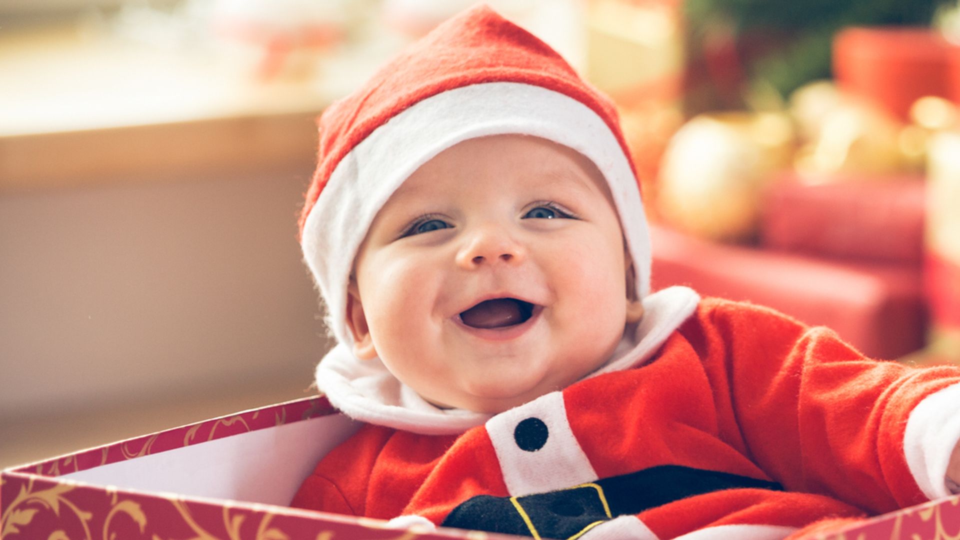 Cute little baby boy in Santa hat sitting inside of the red gift box next to the Christmas tree.