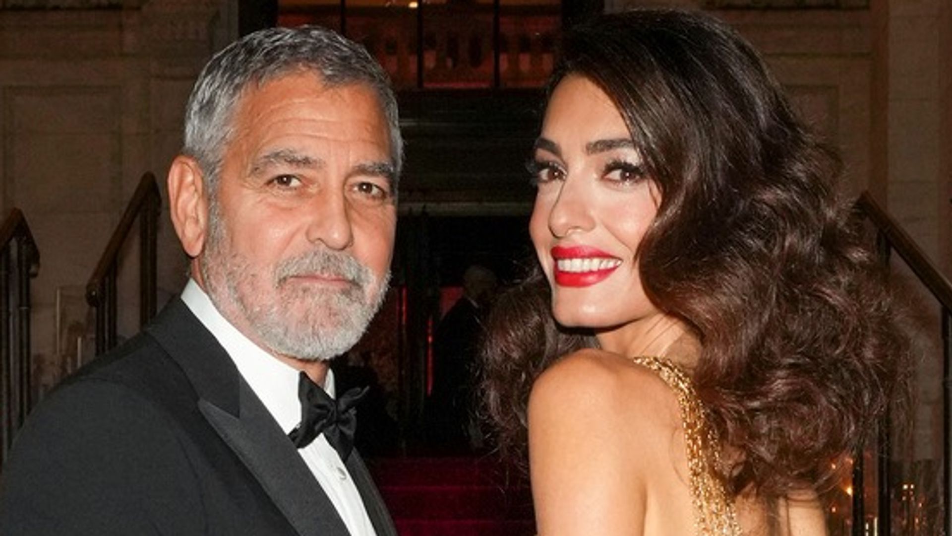 Amal Clooney in a gold dress and George Clooney in a suit as they walk up stairs with a red carpet