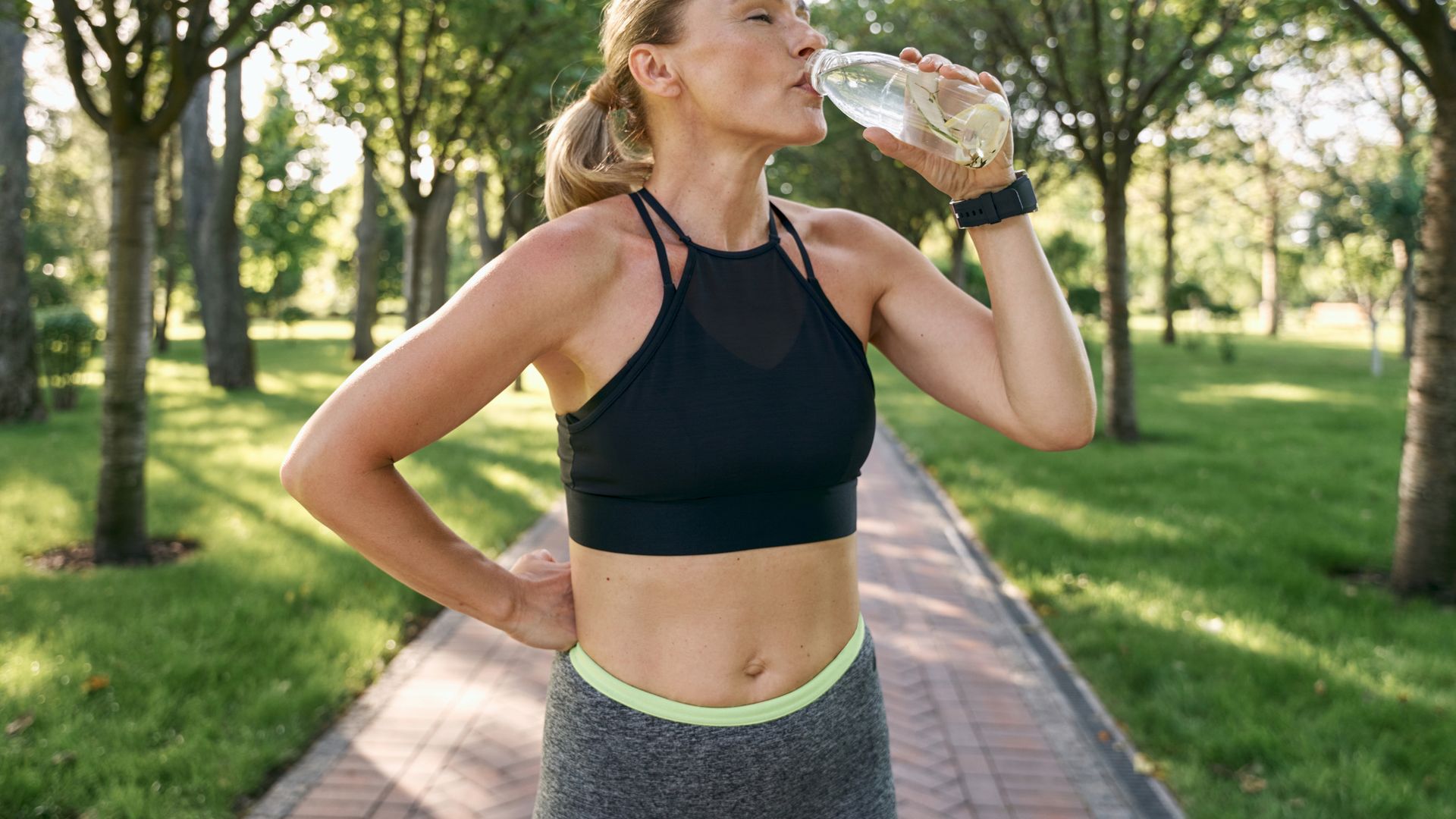 Hydration. Thirsty sporty woman in sportswear drinking water after jogging in a green park on a sunny summer day. Sport, healthy lifestyle concept