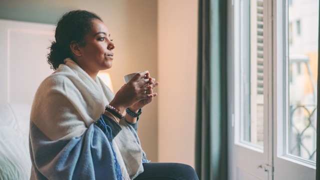 Woman staying home for safety during coronavirus pandemic, drinking hot tea and looking through the window