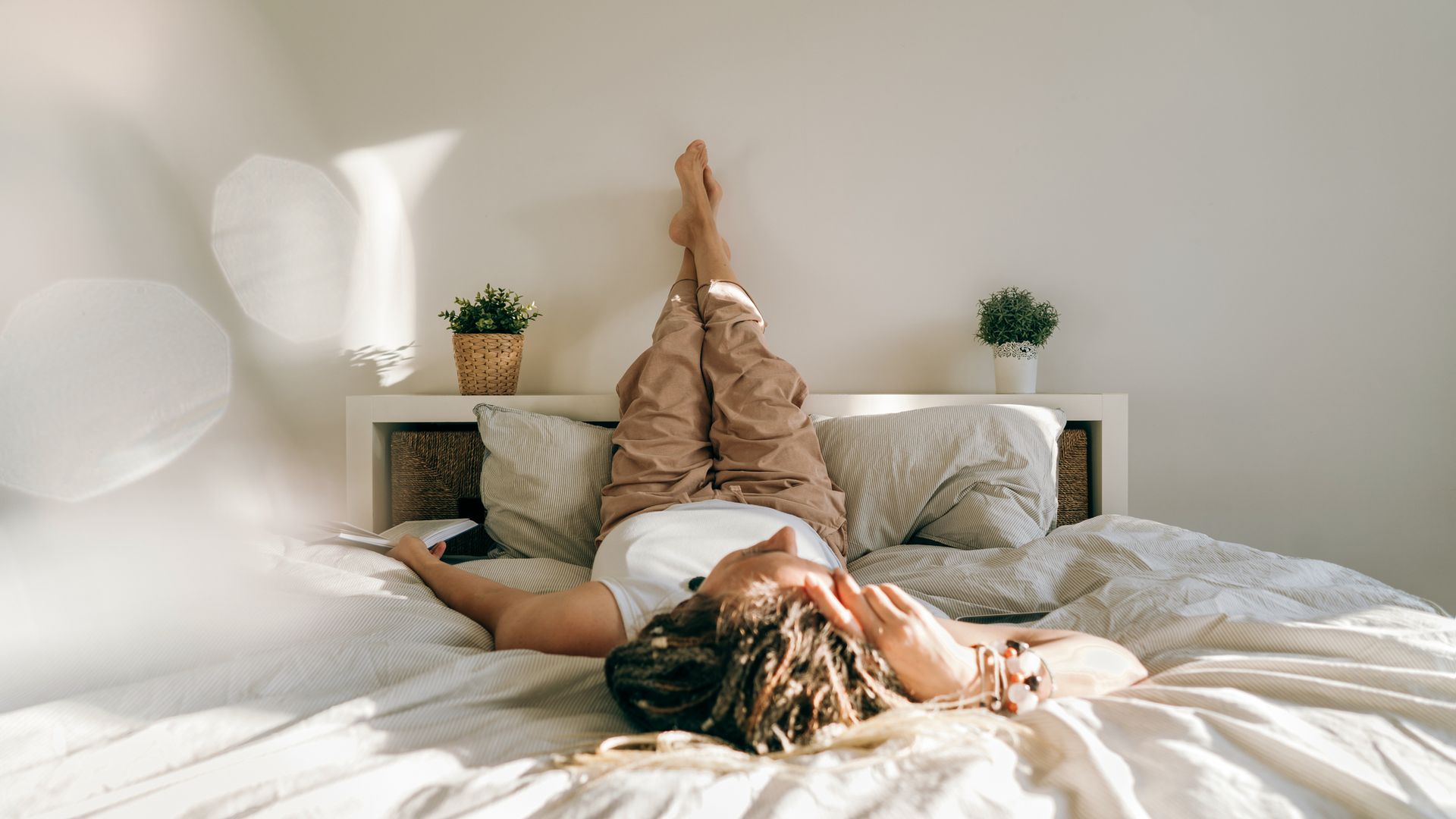 Woman in home clothes book lying on the bed reading. Work at home. Portrait of a dreadlocks braid woman legs up, inverted pose, swelling prevention varicose veins. High angle view