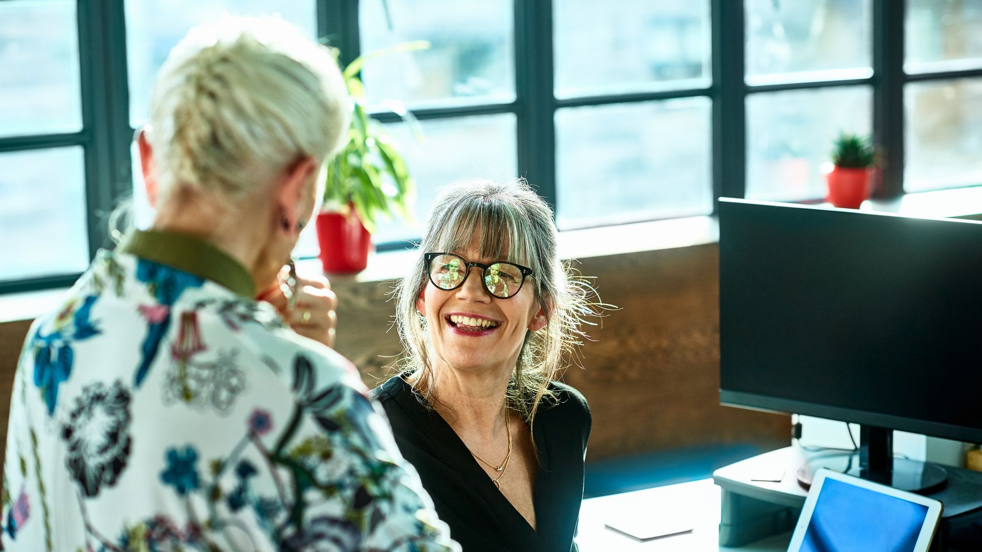 Two friends having conversation at work, woman in her 50s wearing glasses smiling and facing female manager, agreement, support, collaboration
