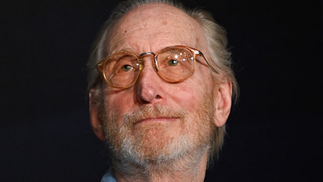 Charles Dance in a blue shirt and glasses looking into the distance on a black background