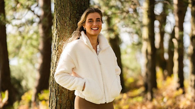 woman in brown leggings and a fluffy white fleece smiling while leaning on a tree