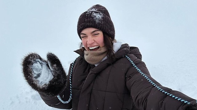 Lily James in the snow wearing white trousers and a brown jacket