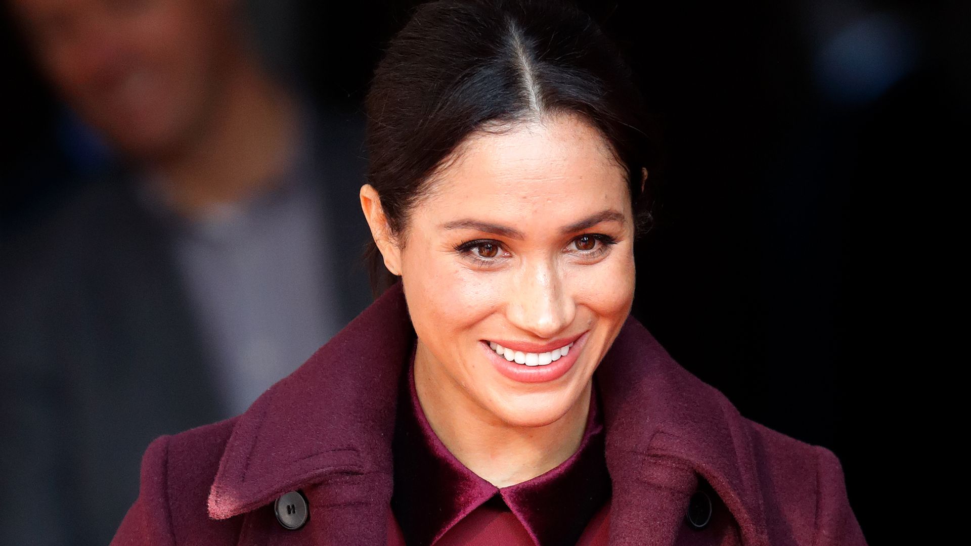 Meghan Markle smiles wearing a red coat