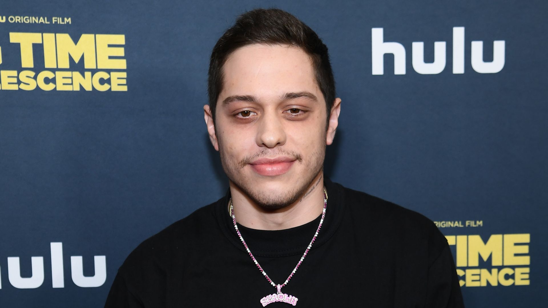 photo of pete davidson wearing black on red carpet with hands folding in front of him