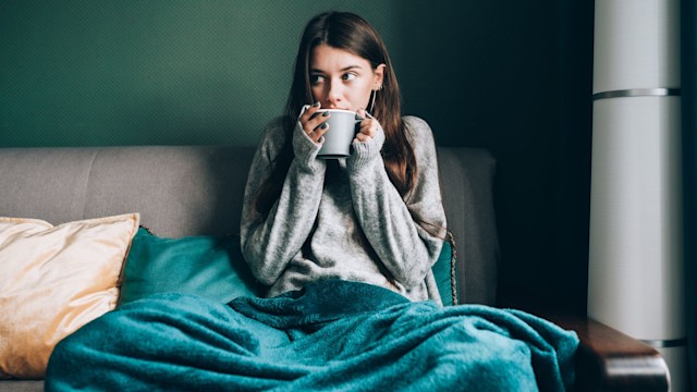 Shivering, frost brunette woman in sweater, wrapped in duvet sit on sofa and drink hot tea