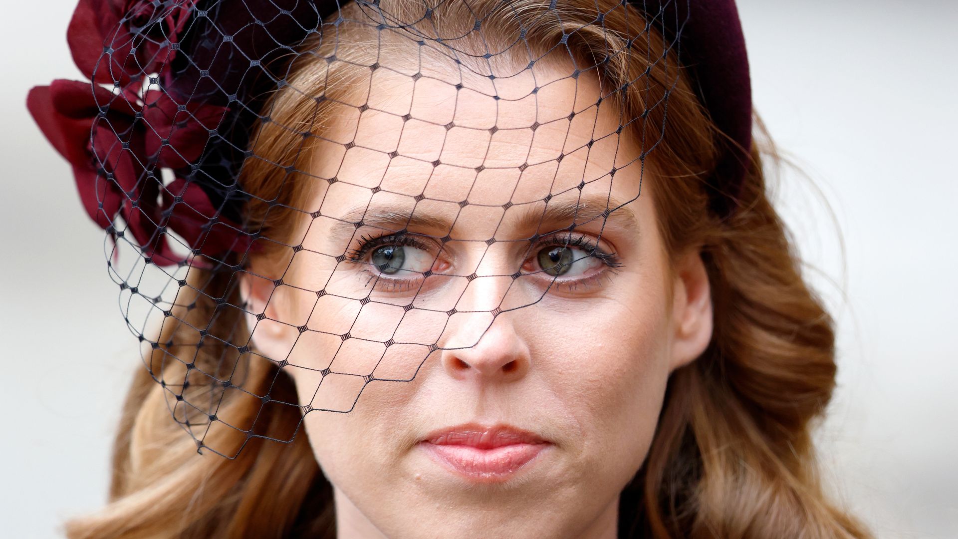 Princess Beatrice looking to the right with a serious expression, wearing a marroon fascinator and navy cardigan