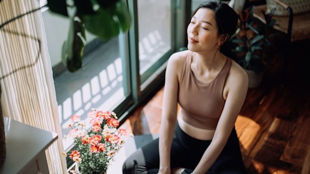 Active young Asian sports woman taking a break after working out at home, sitting on exercise mat taking a deep breath with her eyes closed. Sports and exercise routine. Health, fitness and wellness concept