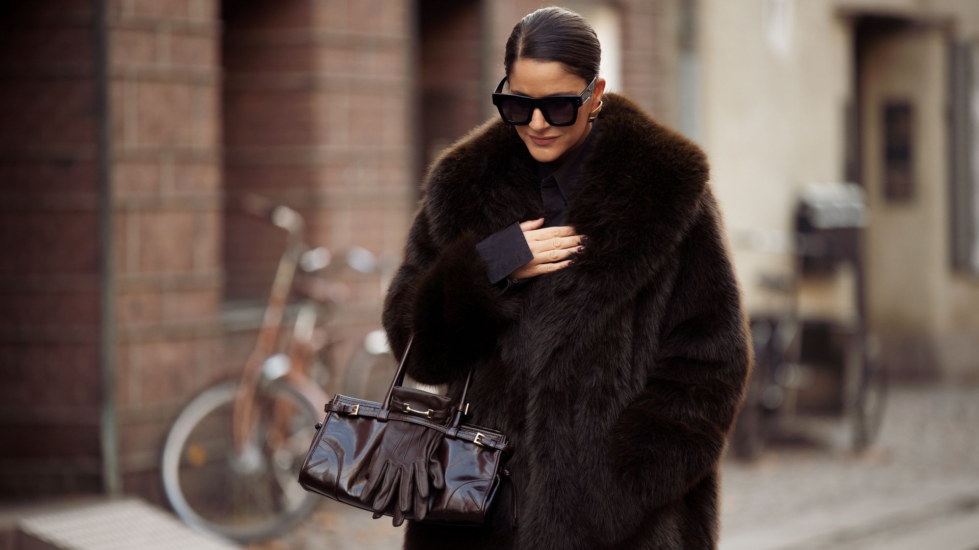 Gili Biegun wears chocolate brown Prada bag, brown leather gloves by Strathberry, black sunglasses by Kaleos, faux fur brown coat by Zara on December 03, 2024 in Copenhagen, 