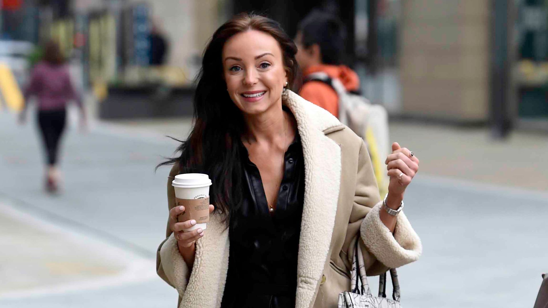 Helen George smiling in a tan coat and black dress while holding a takeaway cup