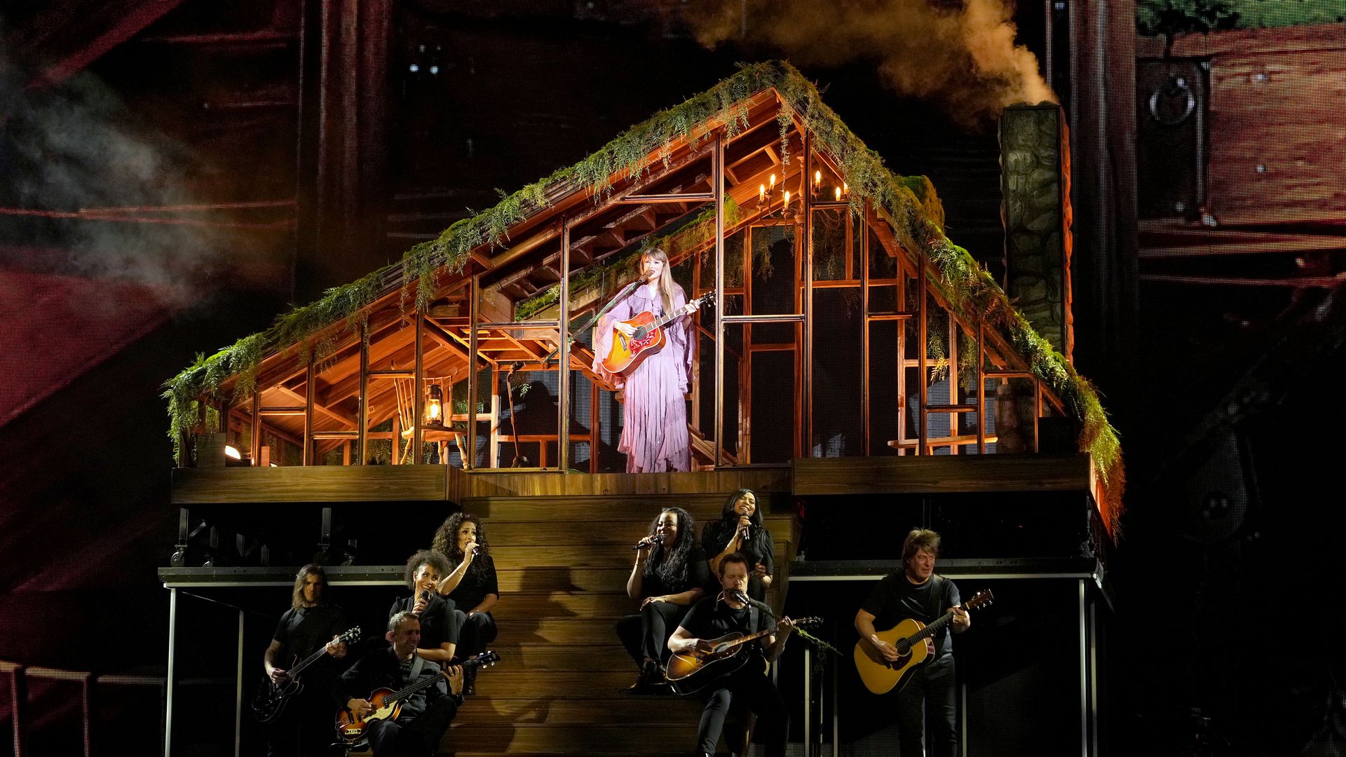 Taylor Swift singing on stage with a guitar surrounded by the set of an overgrown house