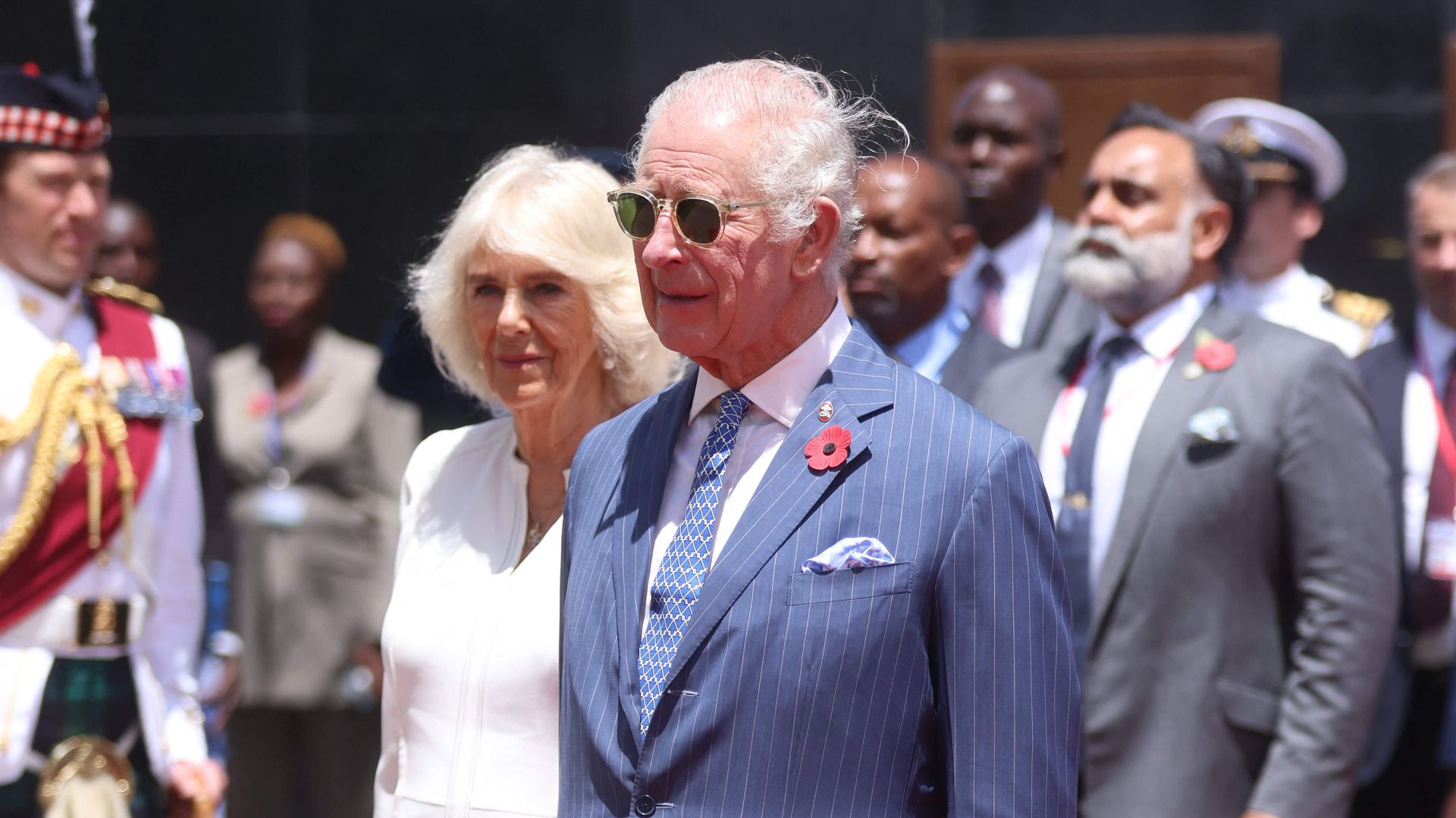 King Charles wearing sunglasses and a blue suit whilst standing next to Queen Camilla