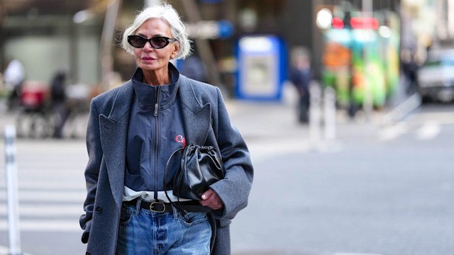 Grece Ghanem wears black sunglasses, dark navy blue white zipped high neck jacket, dark blue double breasted wool coat, shiny black Calvin Klien drawstring leather bag, black leather belt, outside Calvin Klein, during the New York Fashion week Fall/Winter 2025 on February 7, 2025 in New York, New York. 