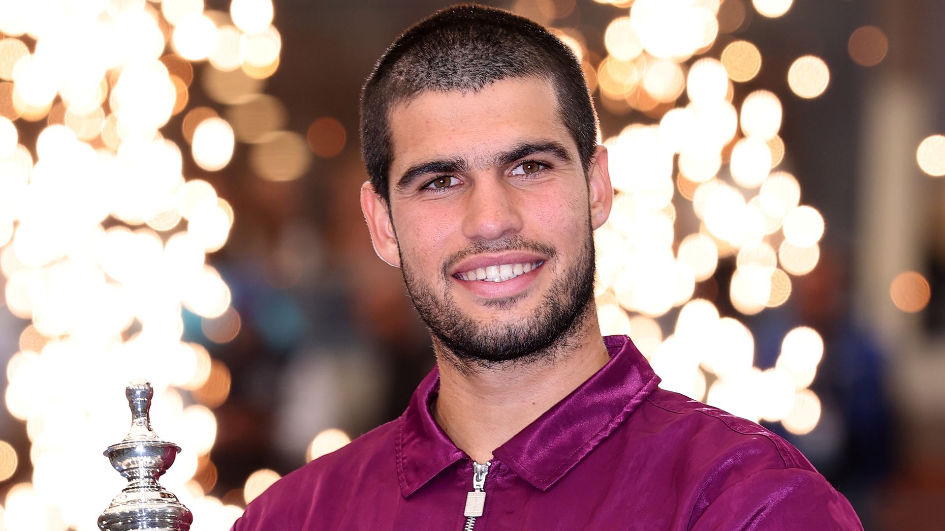 Alcaraz in a purple jumper holding his trophy in front of flares