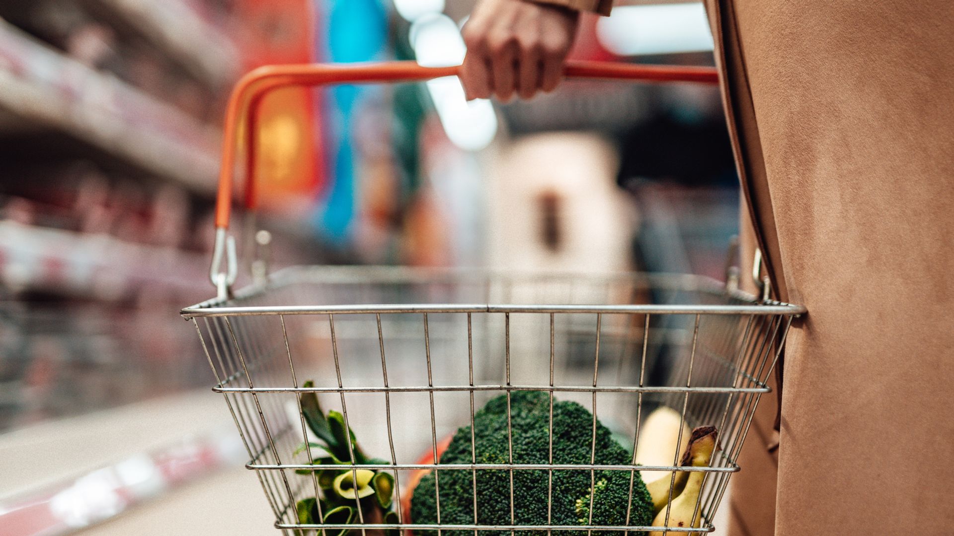 Cropped shot of female hand carrying shopping basket while choosing vegetables at supermarket. Grocery shopping concept.