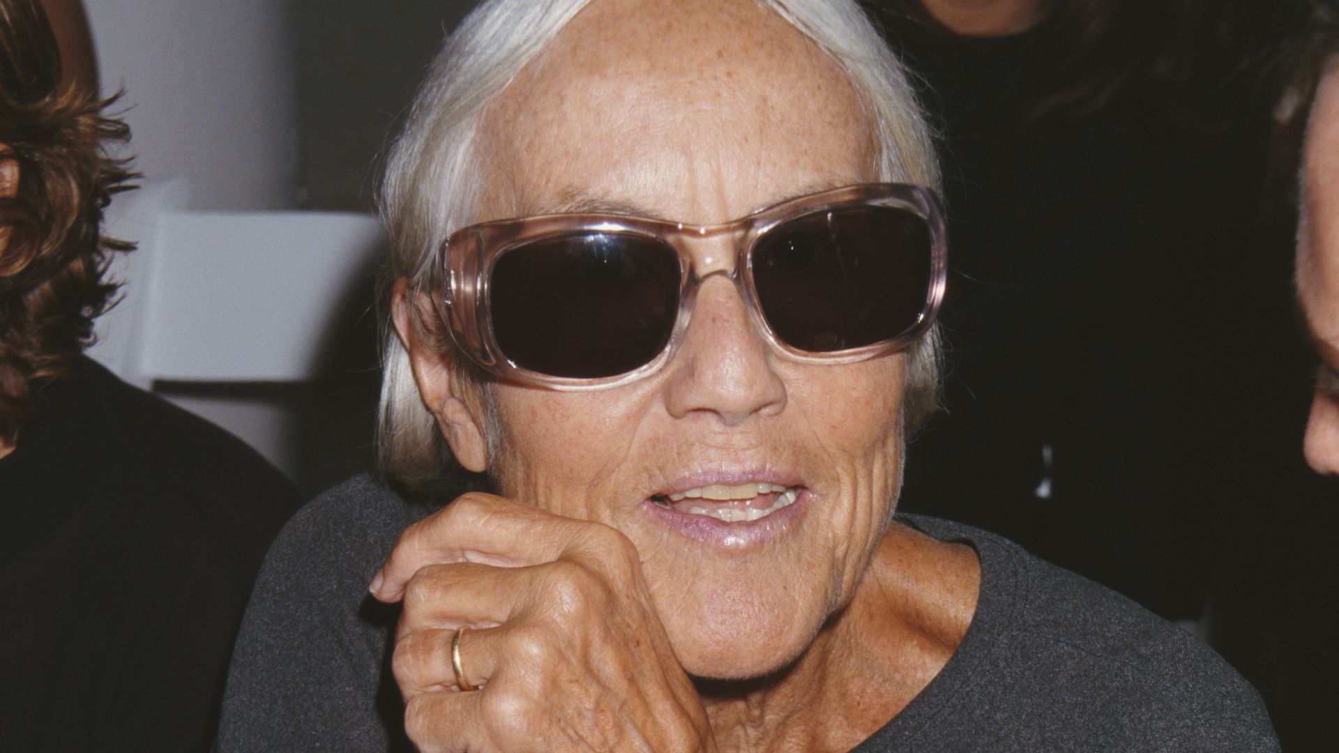 This image shows Polly Mellen, a fashionable older woman with short gray hair, wearing dark sunglasses and a black shirt. She has silver bracelets and watches, exuding a chic, confident vibe.