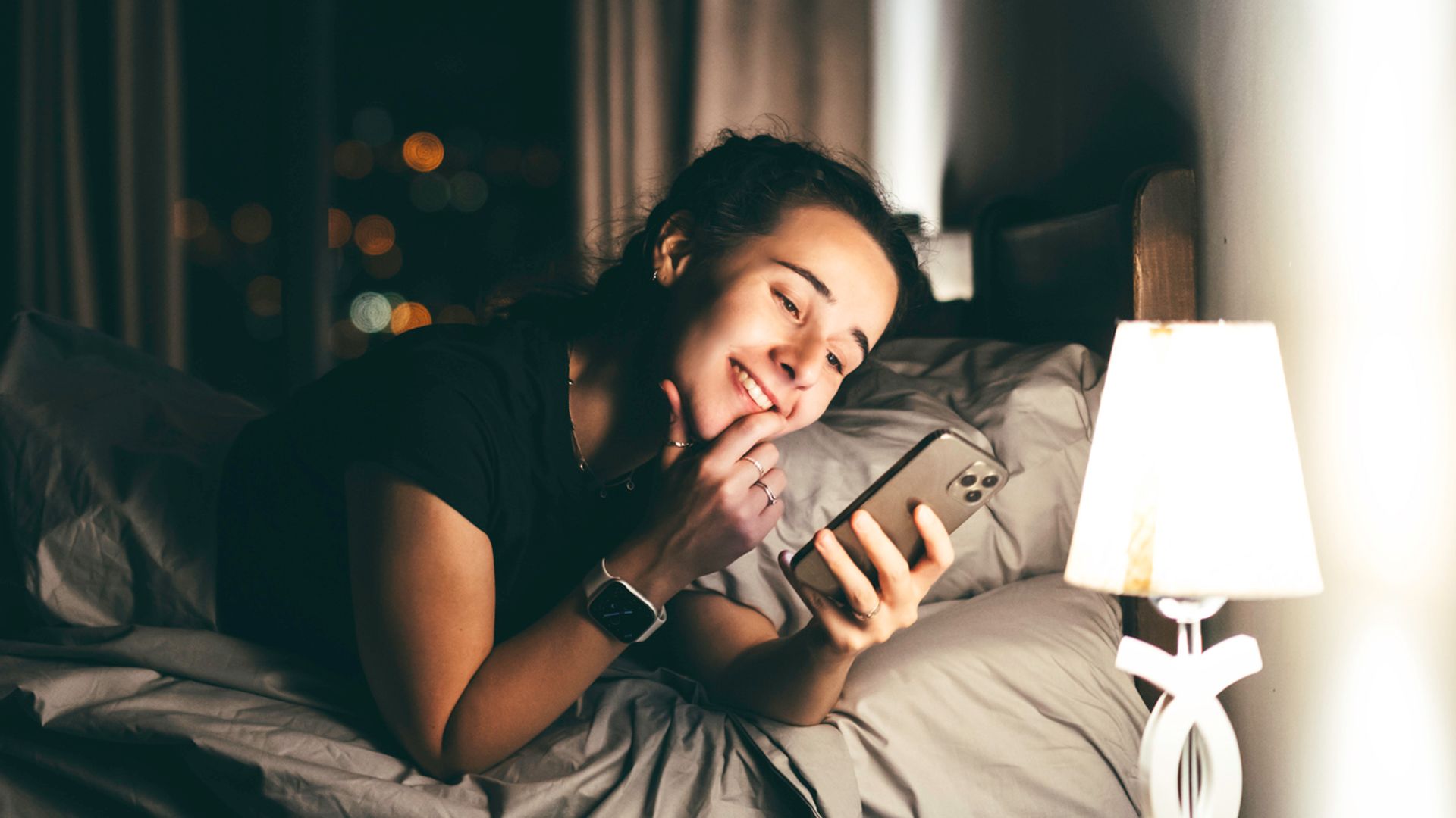 Late night shot of a person lying in bed using the phone to surf the social media before sleep. The woman is happy, chatting with someone she likes, having a long-distance relationships or romance. Gadget addiction, bad habits leading to insomnia, and digital detox. NIght city lights on the background.
