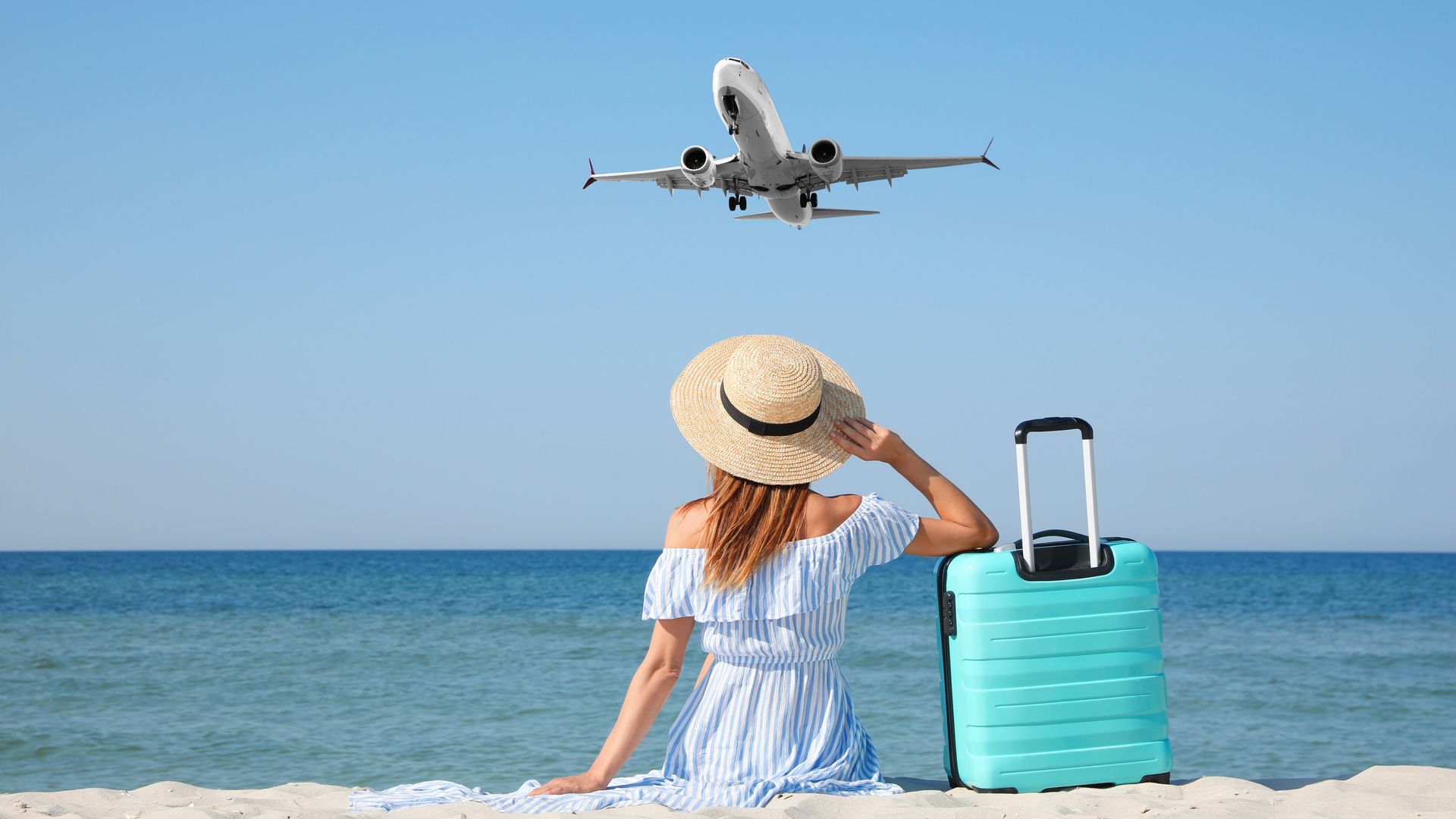 Woman with suitcase on sandy beach looking at airplane flying