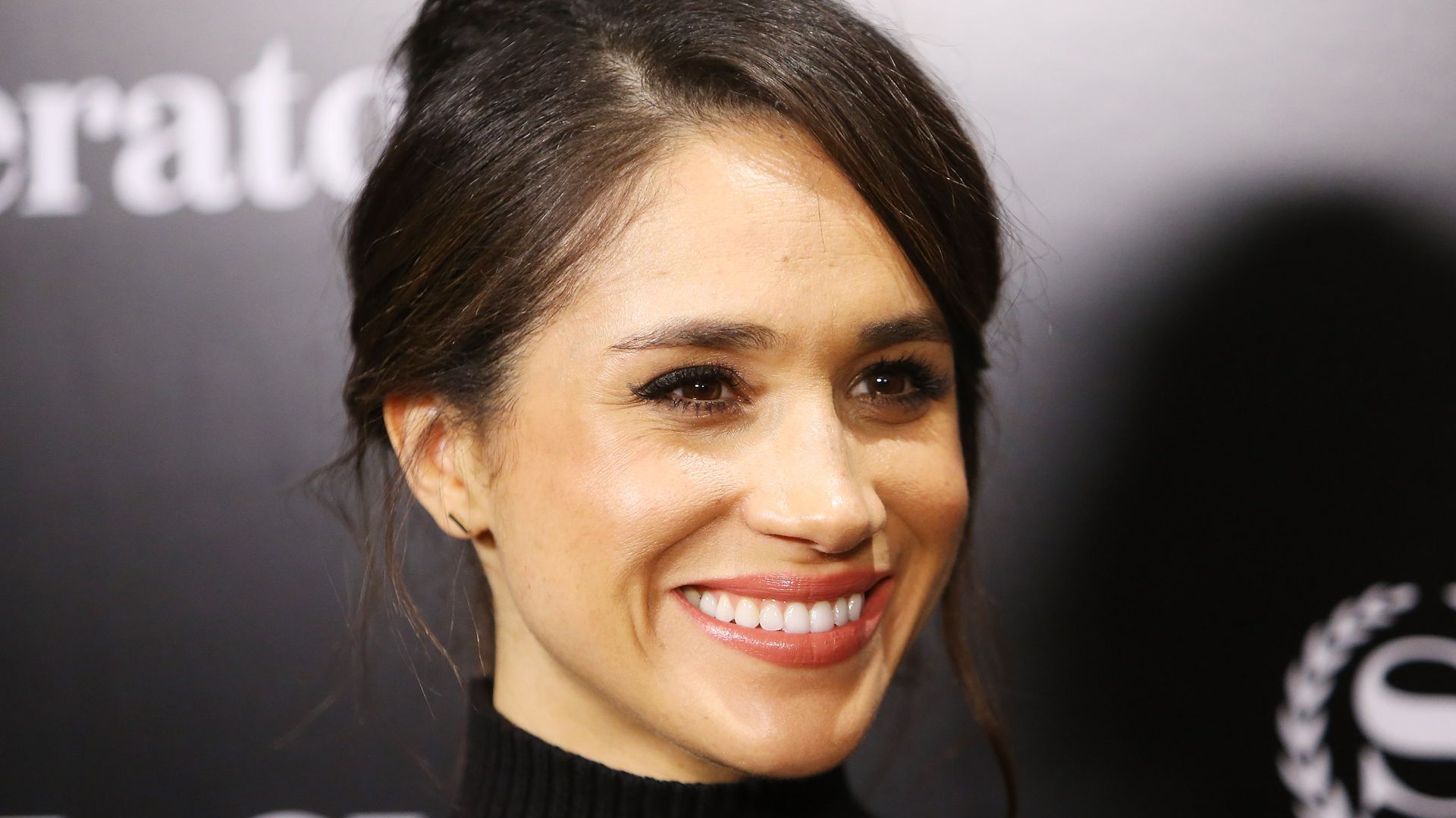Meghan Markle smiling in black sweater with hair up