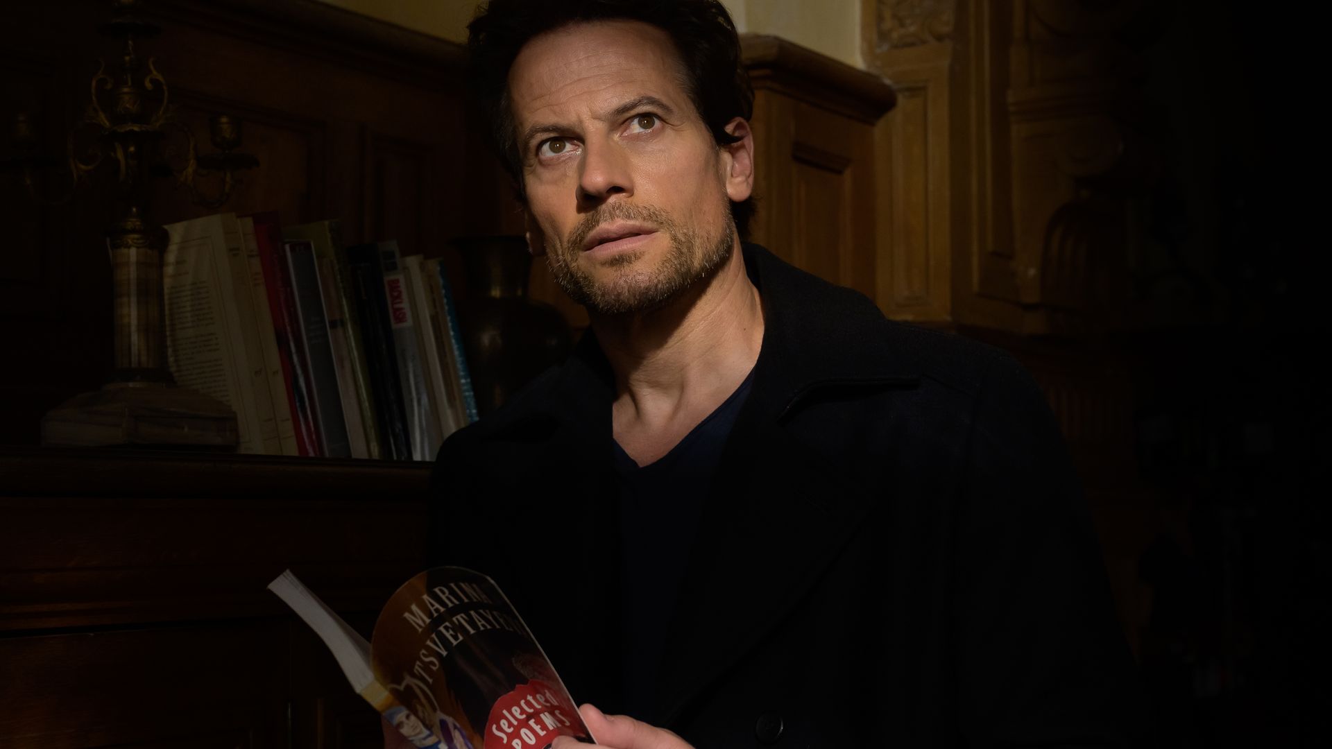 man reading book in dimly-lit room in front of bookshelf