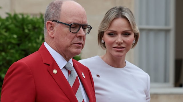 Princess Charlene in white linking arms with Prince Albert in a red jacket