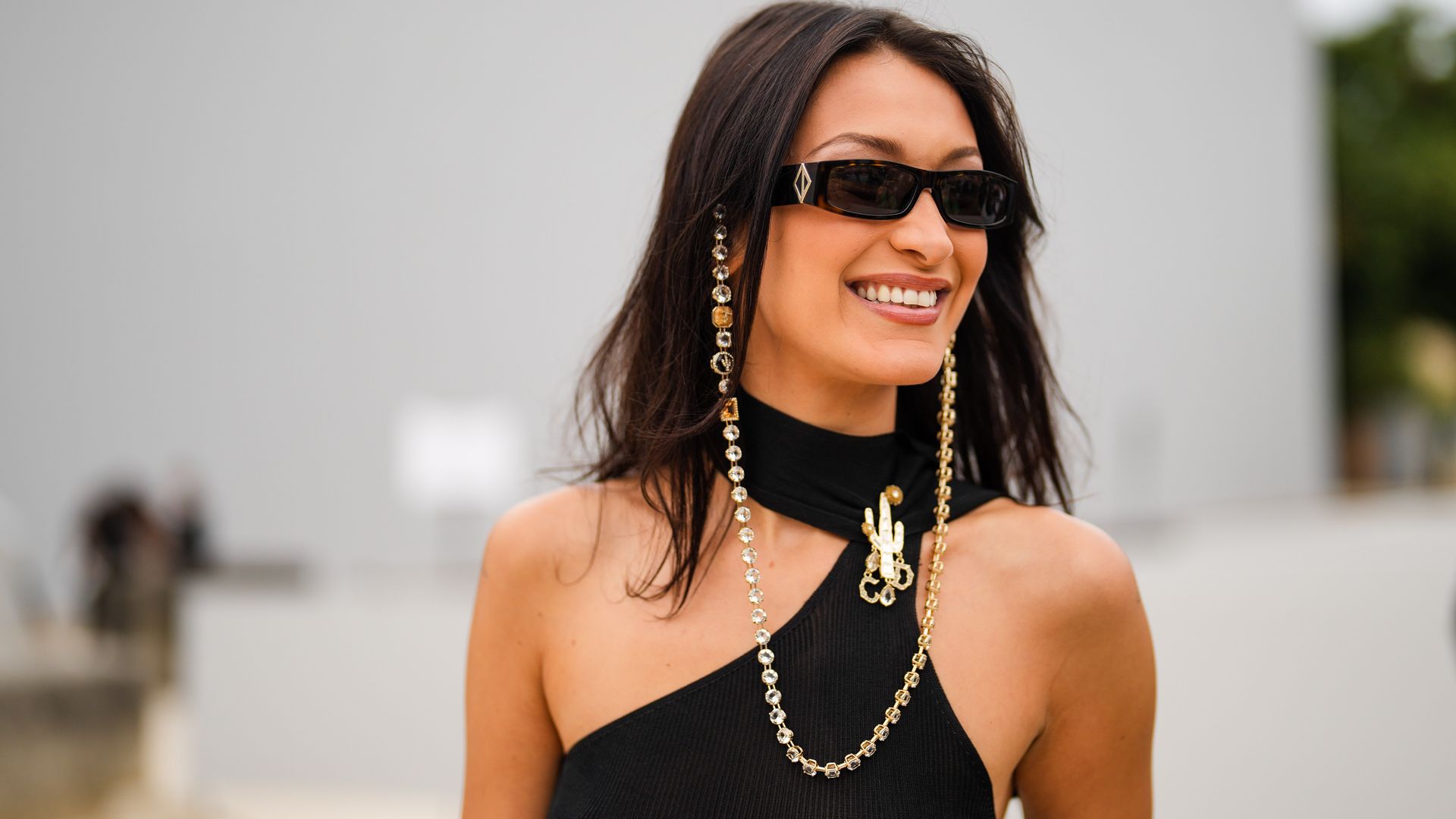 Bella Hadid wears sunglasses with a bejeweled / diamonds chain, a black off-shoulder cropped top with turtleneck, outside Dior, during Paris Fashion Week