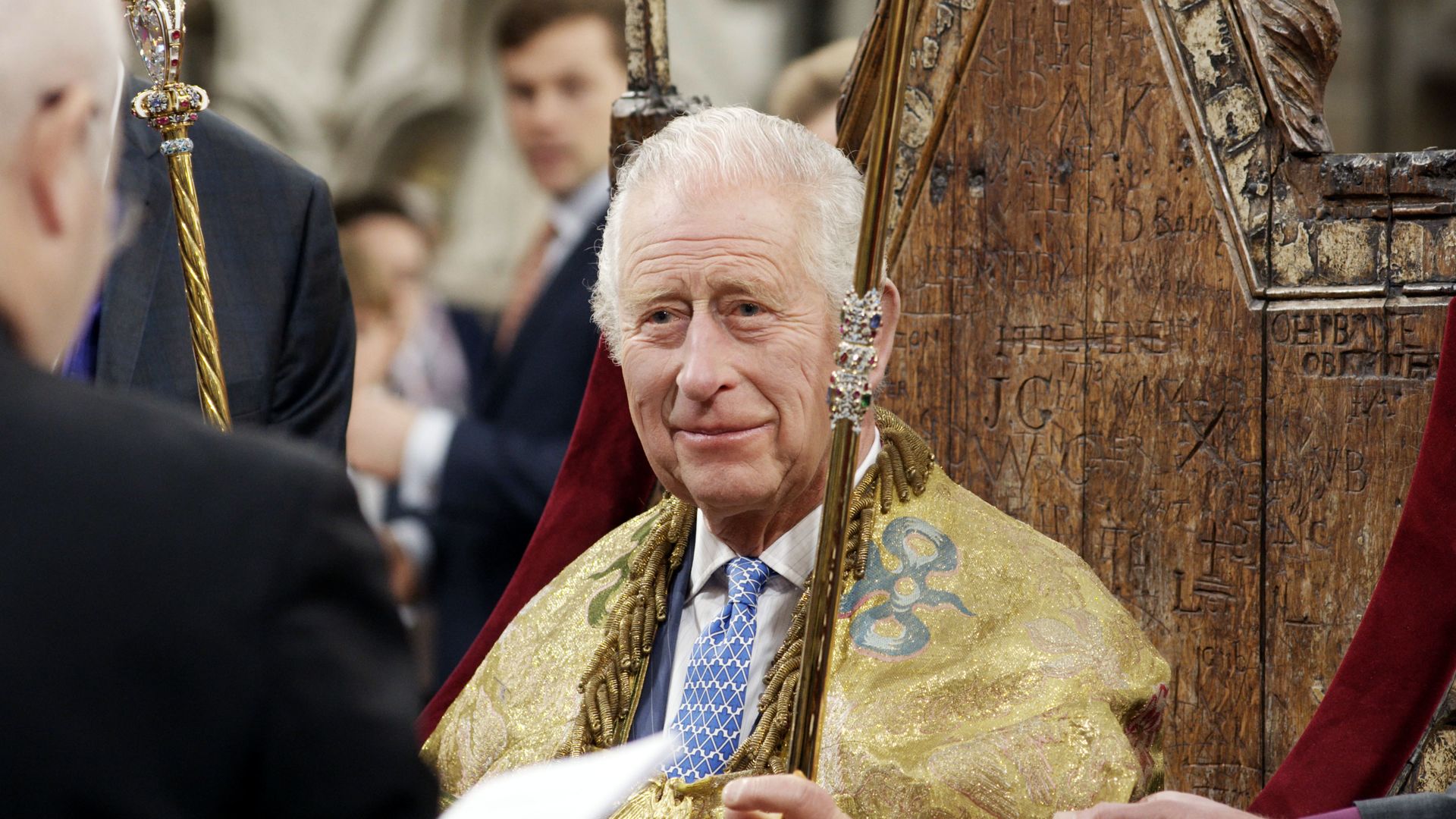 King Charles in a suit and tie and golden robe sat on wooden throne