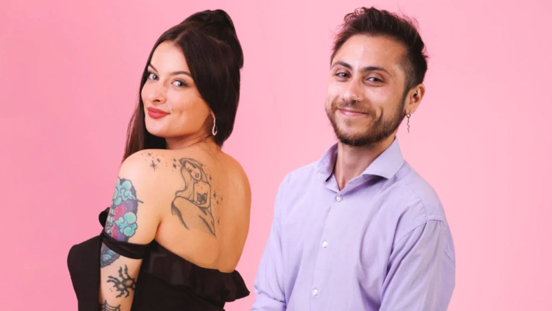 A woman in a black dress and tattoos and a man in a lavendar shirt in front of a pink background