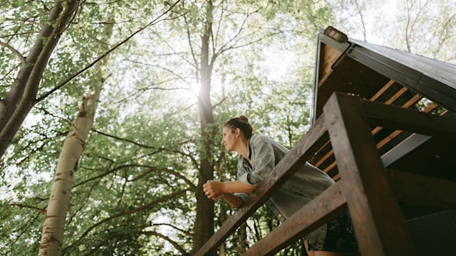 Photo of a young woman enjoying the bright autumn morning on a balcony of a cabin in the woods; weekend getaway in nature, far from the city hustle.