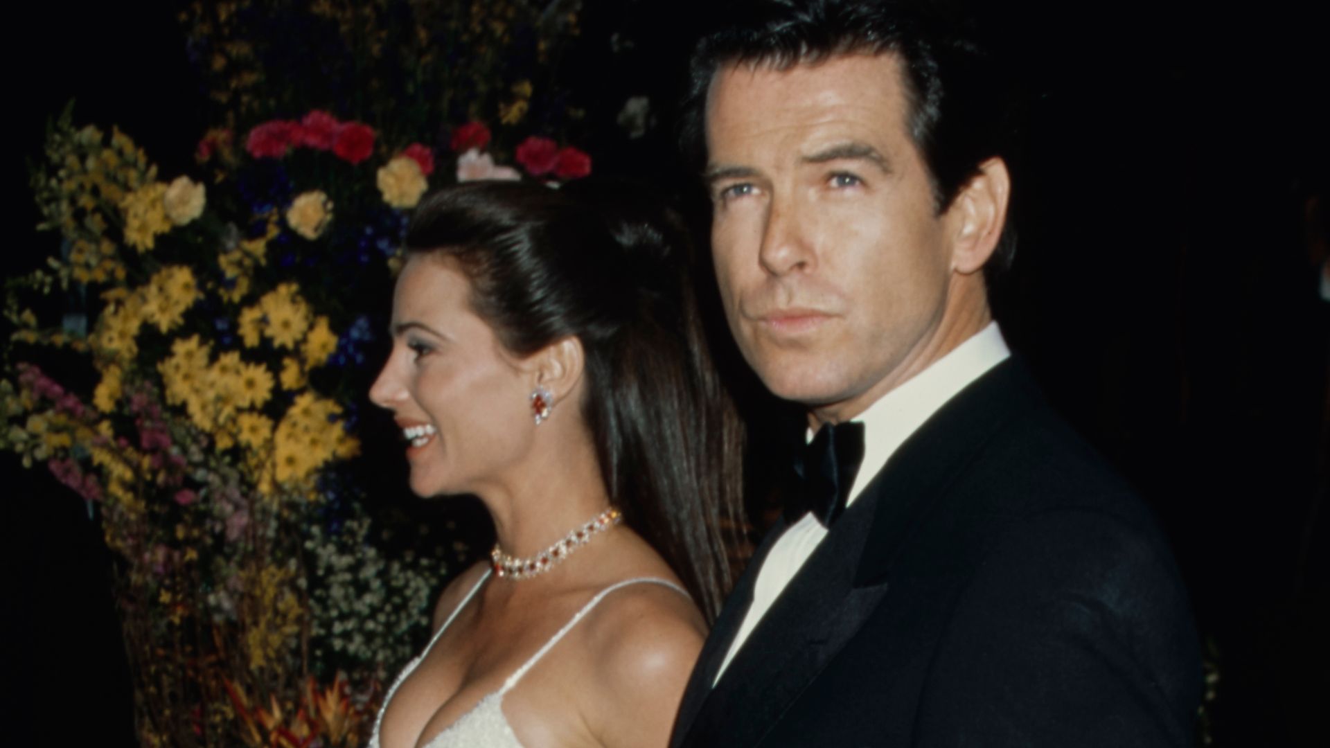 Keely Shaye Smith in a white sparkly dress and Pierce Brosnan in a suit with flowers in the background