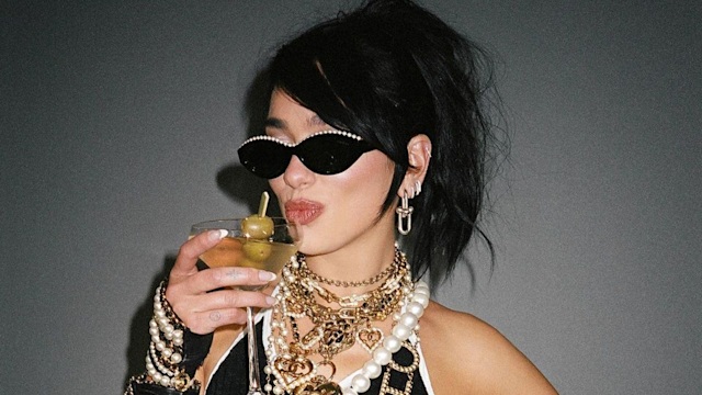 Dua Lipa drinking a martini and wearing layered gold and pearl Chanel necklaces and a monochrome crop top  