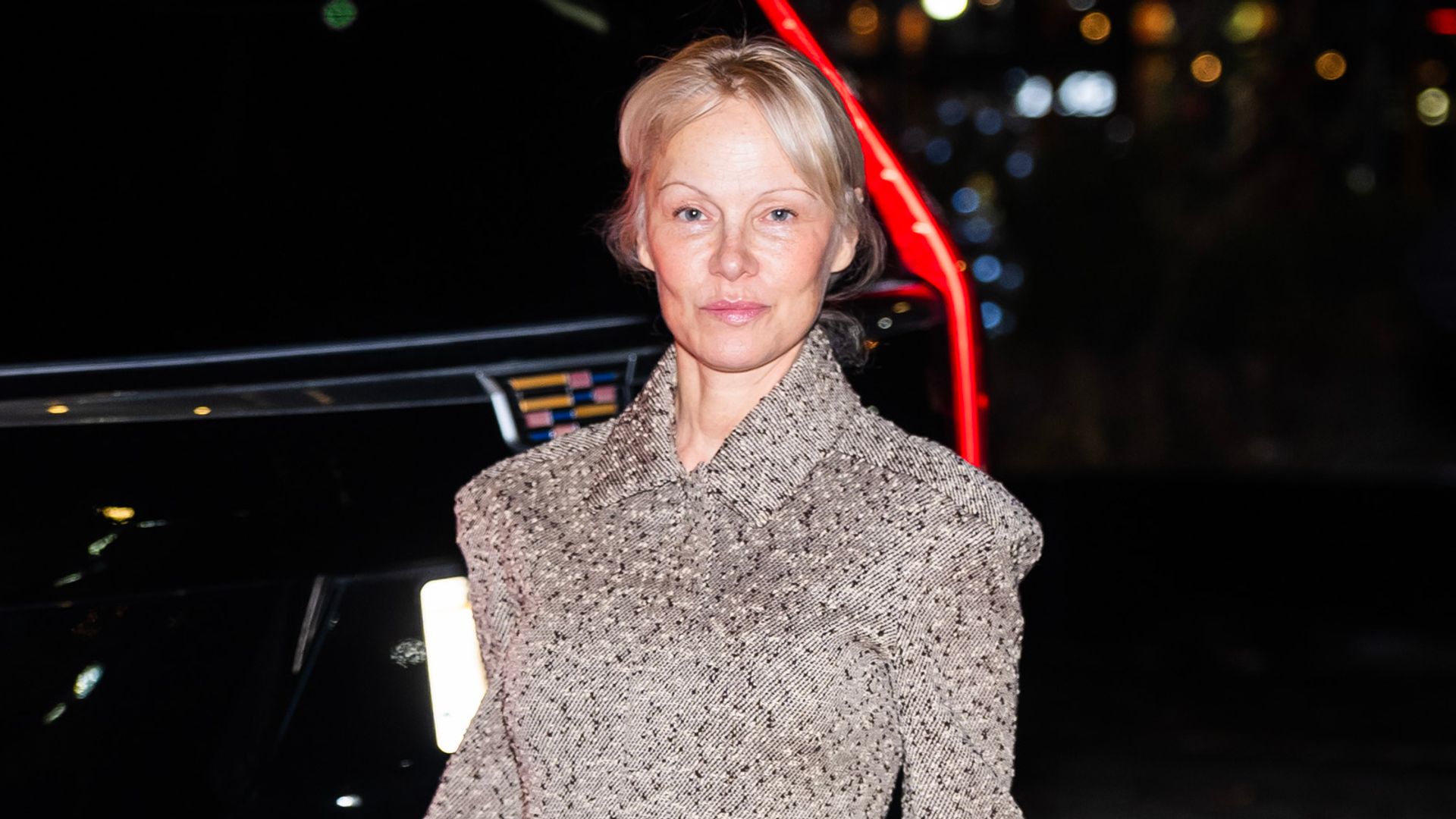 Pamela Anderson walks on a cobblestone street at night in a sophisticated beige tweed Tory Burch ensemble, accentuating her natural glow. Her rich brown knee-high boots add a sleek, modern edge to the tailored look.