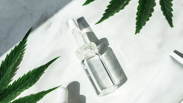 Oil in a frosted and transparent glass jars with a pipette, green leaves of marijuana on a white background with place for text