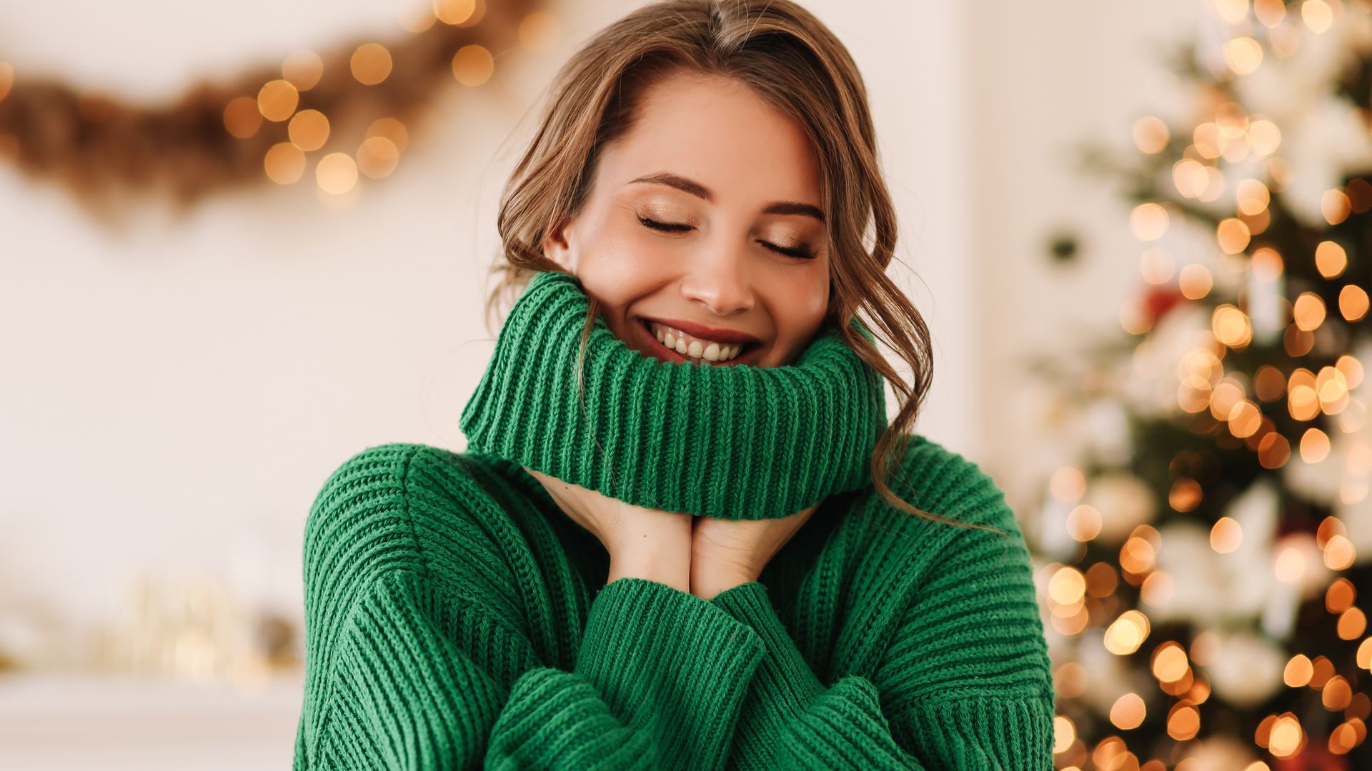 Portrait of a funny emotional cheerful young woman with horns on her head in a green cozy warm sweater laughing having fun holding and eating a lollipop and ginger cookies on Christmas day on the background of a decorated Christmas tree at home in winter on New Year's Eve