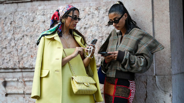 MILAN, ITALY - SEPTEMBER 21: Melanie Darmon (R) wears a gold hair clip, black sunglasses, gold earrings, a brown with blue and red checkered print pattern oversized jacket, a red / black / white embroidered wool pattern knees skirt, ; Gabriella Berdugo (L