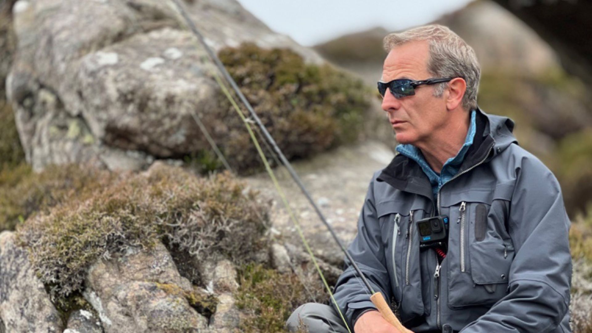 Robson Green with a fishing rod sitting on some rocks