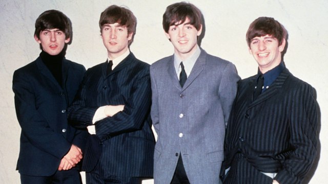 The Beatles stand shoulder to shoulder in tailored suits—dark pinstripes, gray wool, and slim lapels. It’s early Beatlemania, and they look polished, poised, and ready to take over the world.