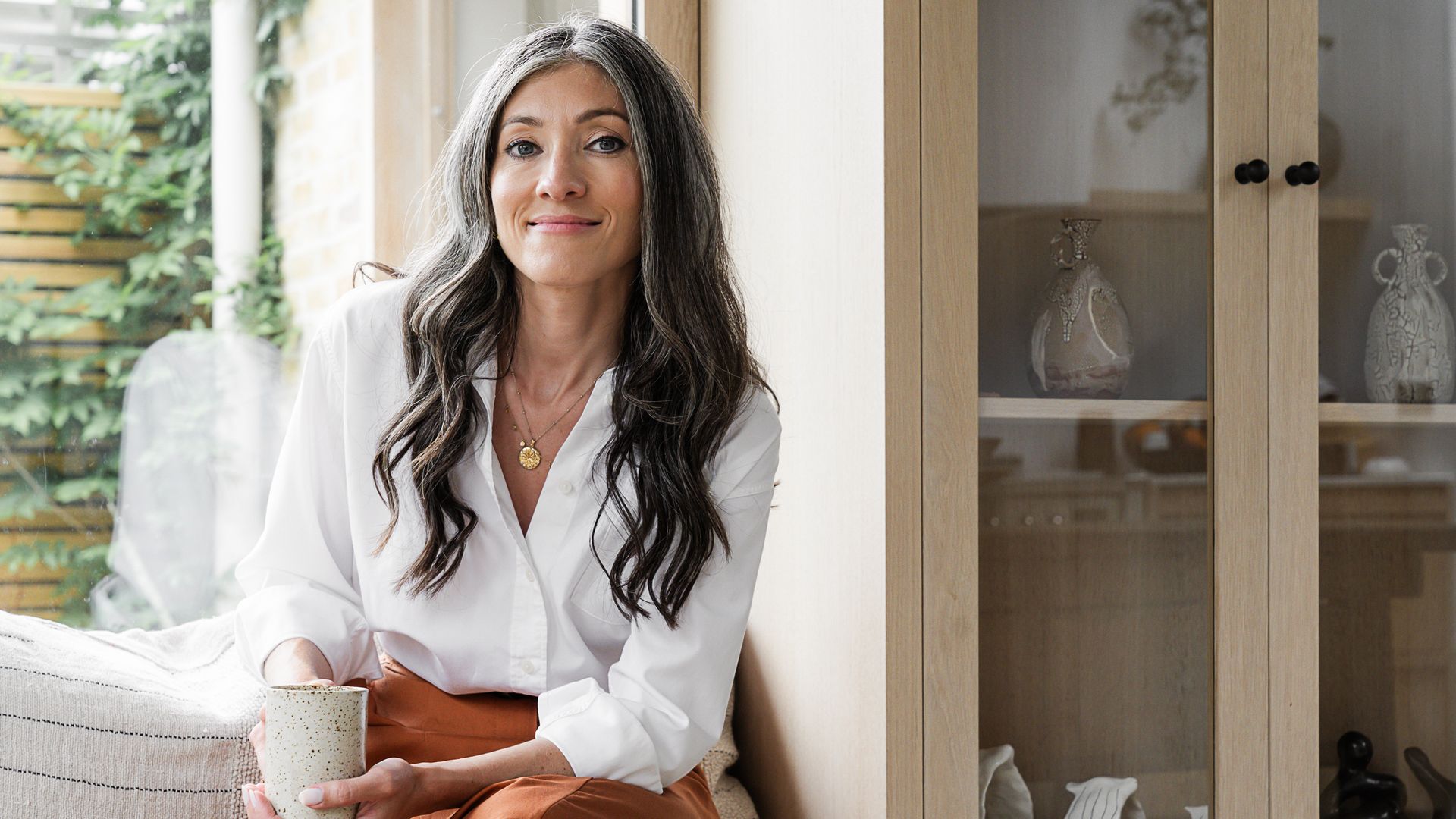 woman with grey hair smiling with a hot drink in a white shirt 