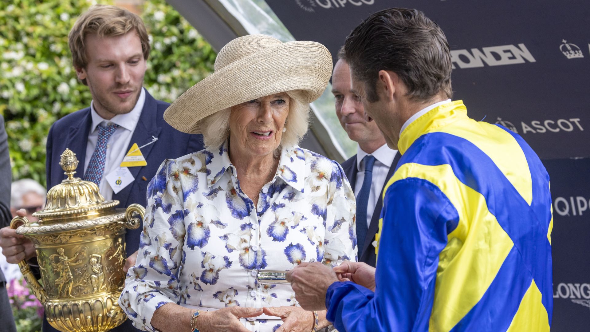 Queen Camilla talking to a jockey - a man behind her holds a golden trophy