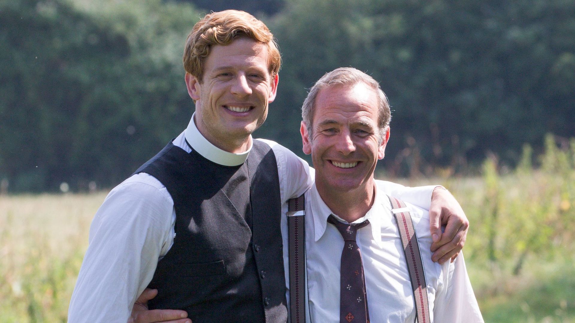 man wearing vicar's collar next to man in suit standing in field