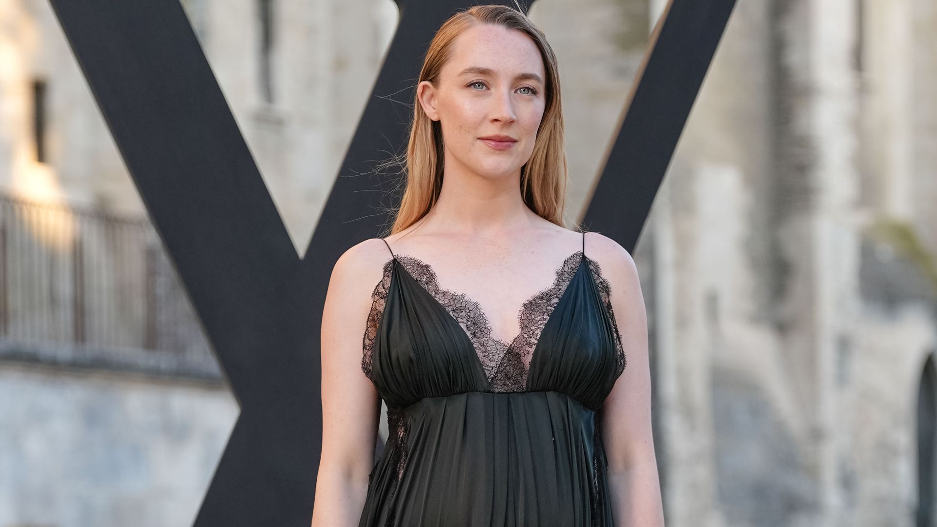 Saoirse Ronan wears a black pleated low-neck dress with dark green hue featuring delicate lace trim and thin straps. Hair is styled straight and parted in the center.