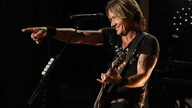Keith Urban facing left performing on stage with a guitar and pointing out at the crowd
