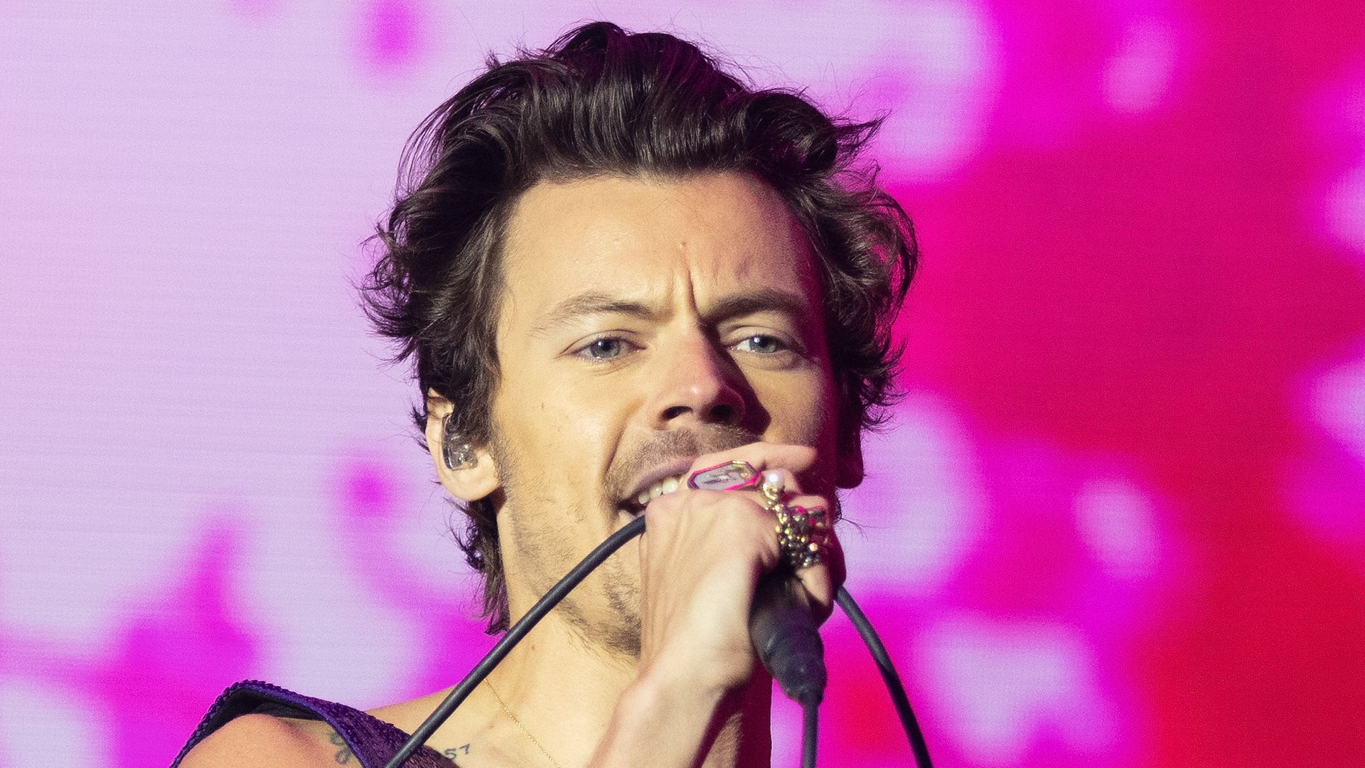 Harry Styles performs on stage, singing into a microphone while wearing a sparkly purple bodysuit