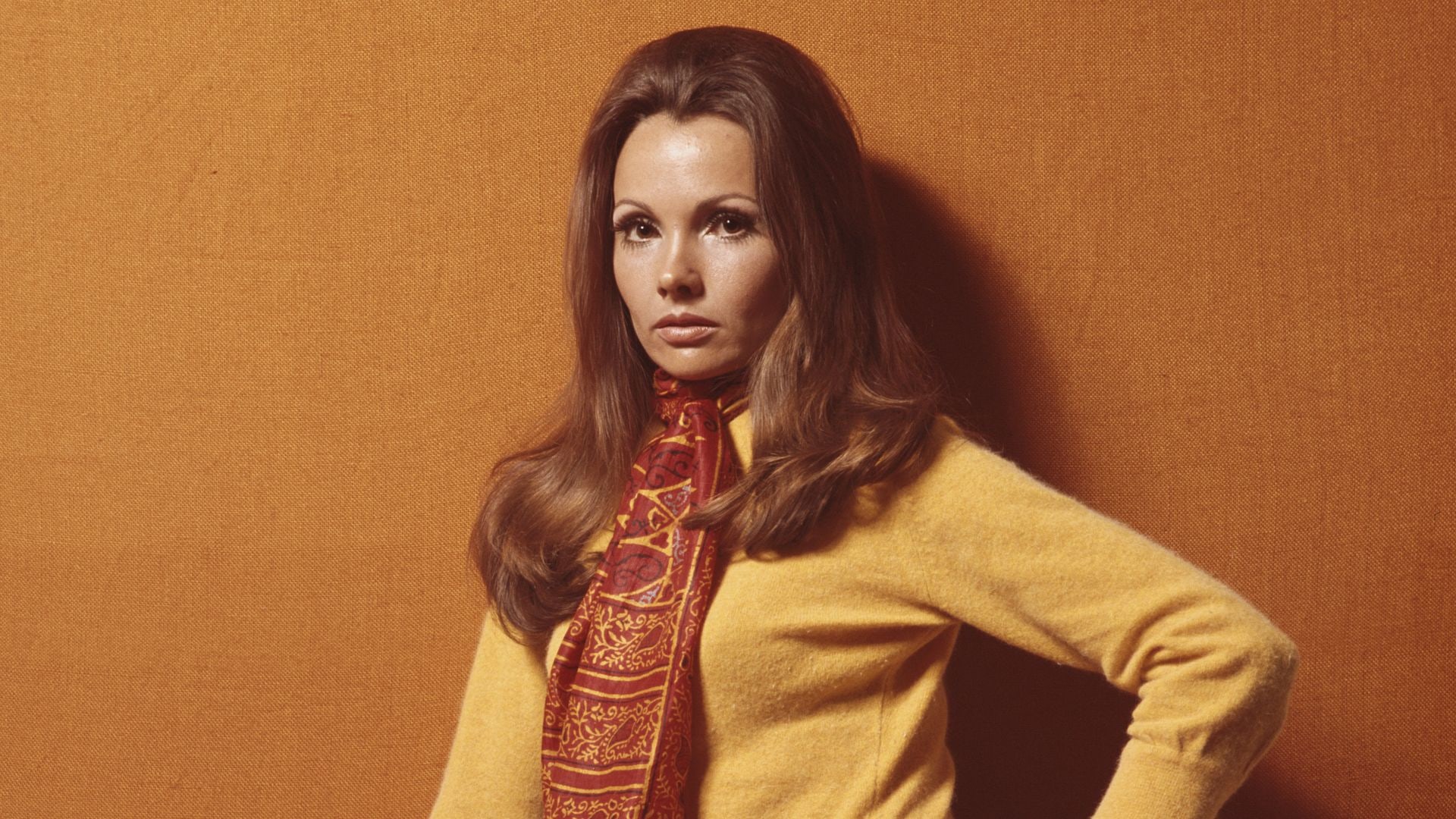 A young woman poses in a purple pleated miniskirt with a yellow top and an orange patterned scarf, circa 1970. 