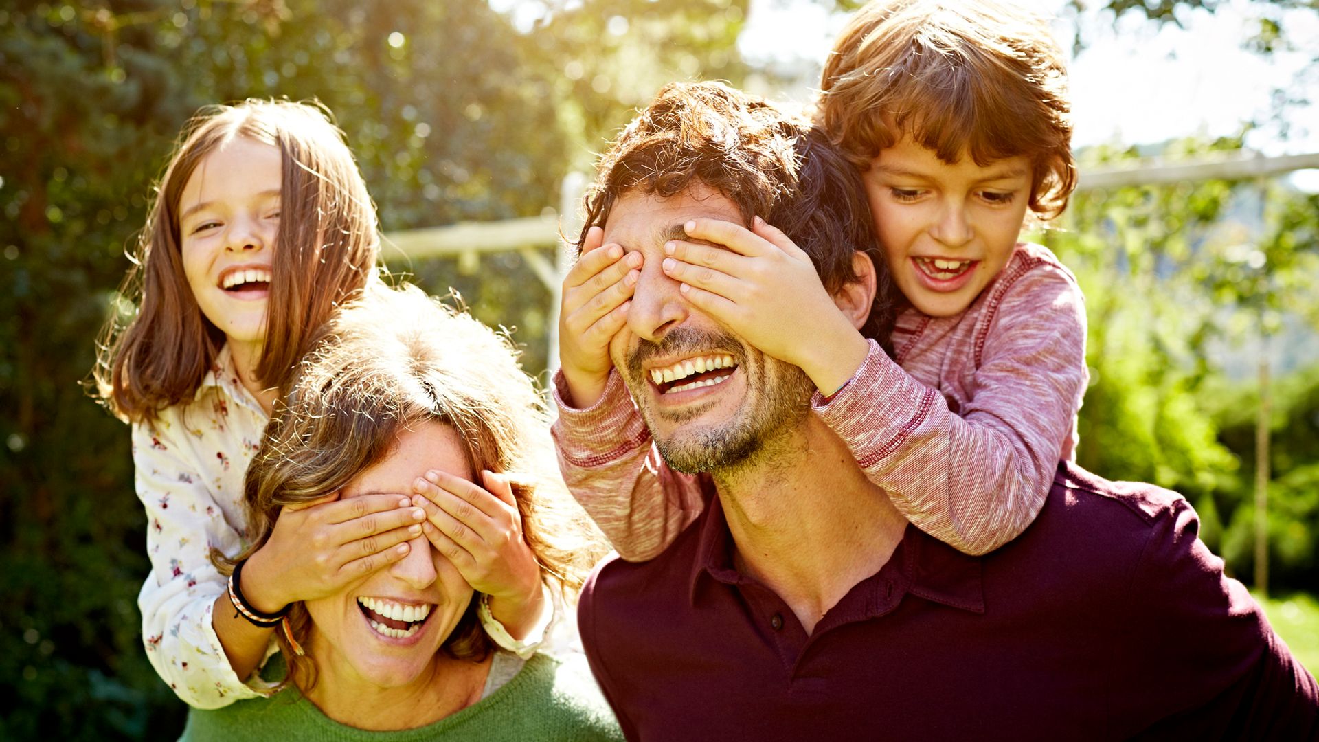 Playful children covering eyes of parents while enjoying piggyback ride in park