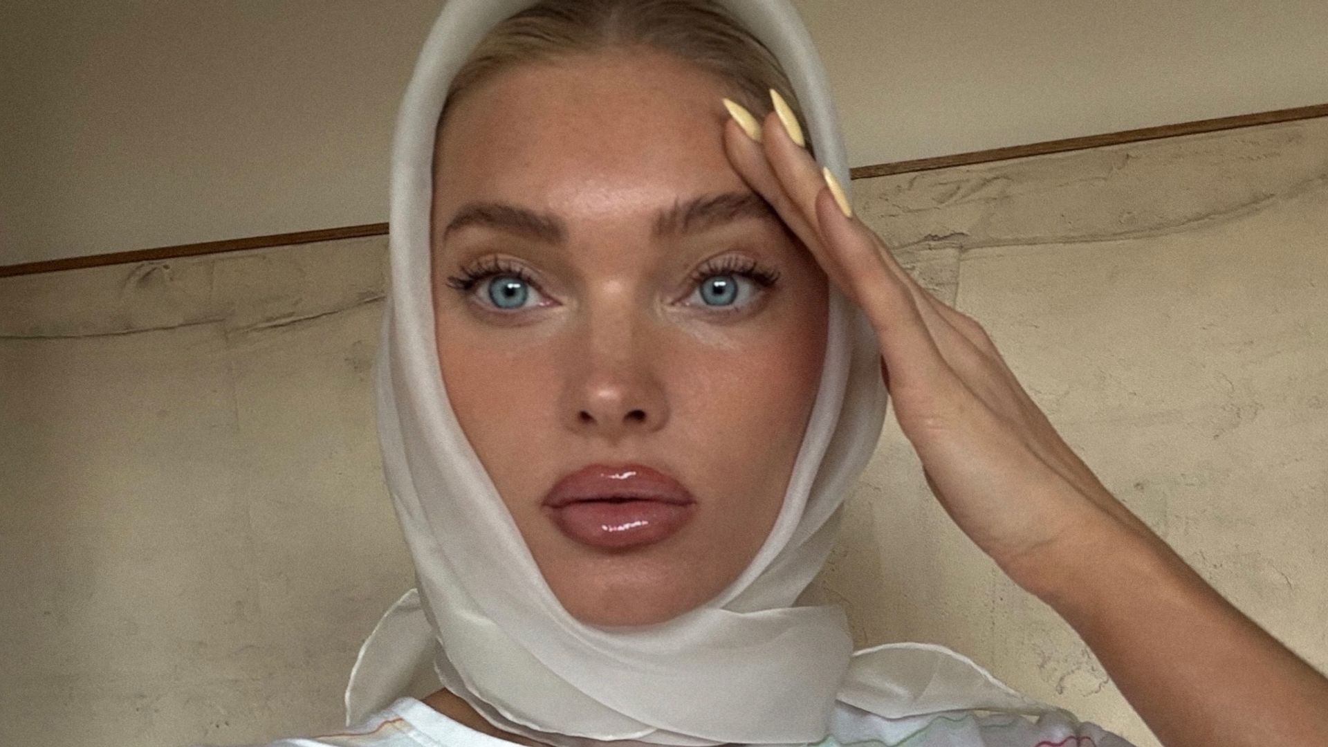 The image presents a close-up portrait of Elsa Hosk draped in a creamy white headscarf, exuding an ethereal glow. Her piercing blue eyes, framed by softly feathered brows and delicately lifted lashes, draw the viewer into a moment of quiet intensity. Her skin appears dewy and sculpted, with a luminous finish that plays with light in the most flattering way. A glassy, neutral lip enhances the natural curvature of her mouth, while a well-placed highlighter on the high points of her face creates an effortless sculptural effect. The touch that elevates this entire look? A minimalist yet on-trend manicure in a buttery yellow hue.