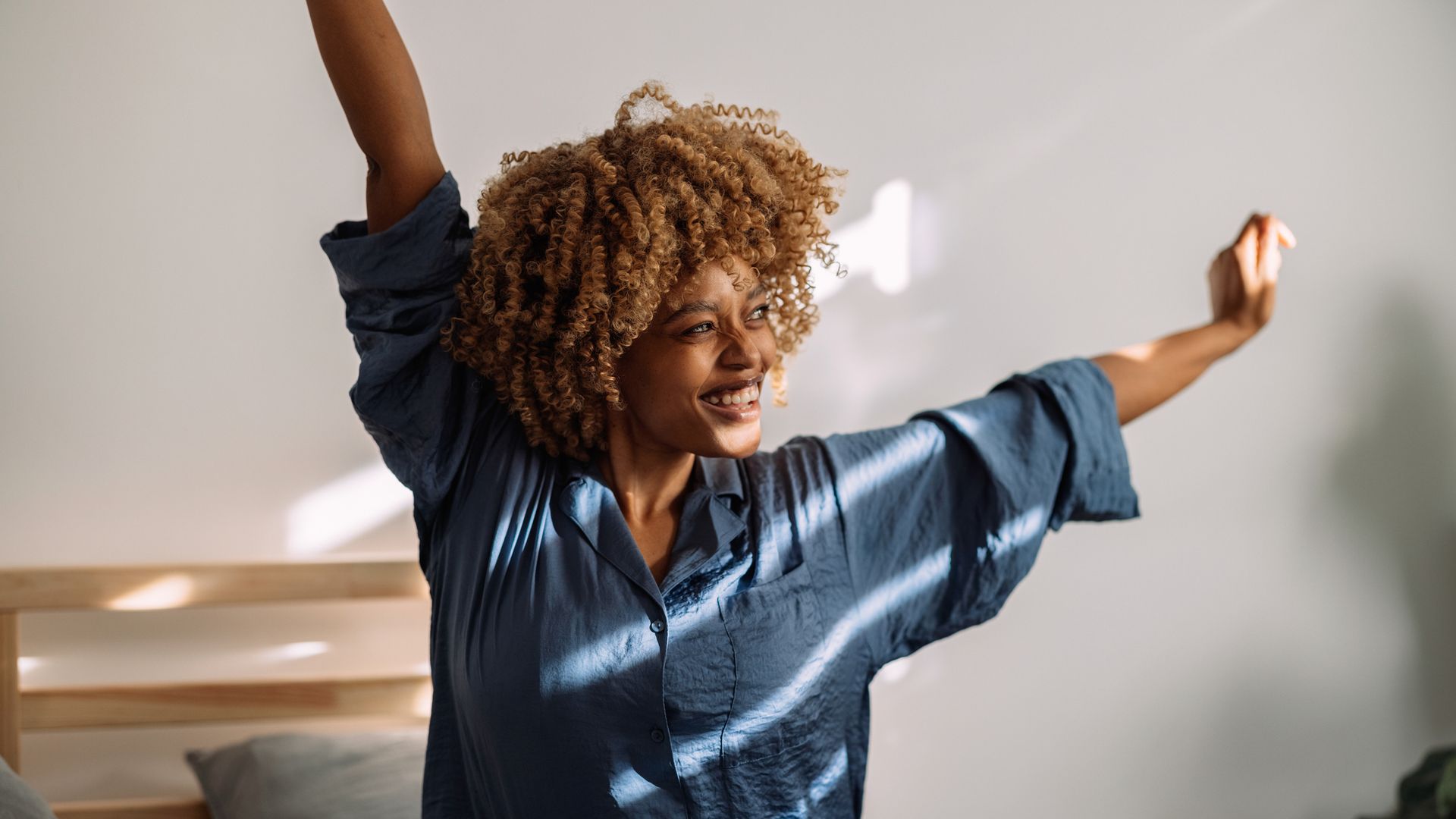 Beautiful woman sitting in her bed in silk pajamas. She is stretching with her arms up in the air and smiling. Sun rays are coming in through the blinds.